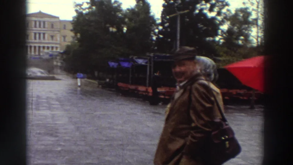 Close-up of a smiling man in a brown coat and dark cap looking back at the camera in a Greek city square circa 1976, with a red umbrella at frame right.