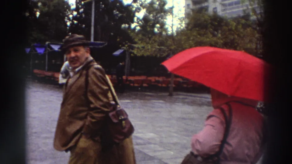 Man in brown coat and hat looking at the camera while a woman in a pink coat holds a large red umbrella in the foreground, Greek city square circa 1976.