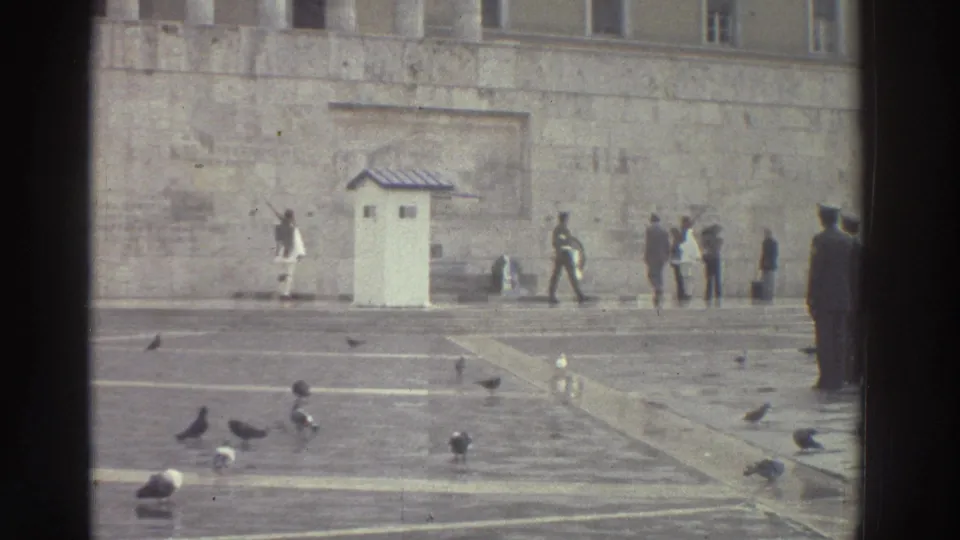 Uniformed ceremonial guards beside a white sentry booth against a large stone wall facade, with pigeons on wet paving stones and civilian bystanders, Greece circa 1976.