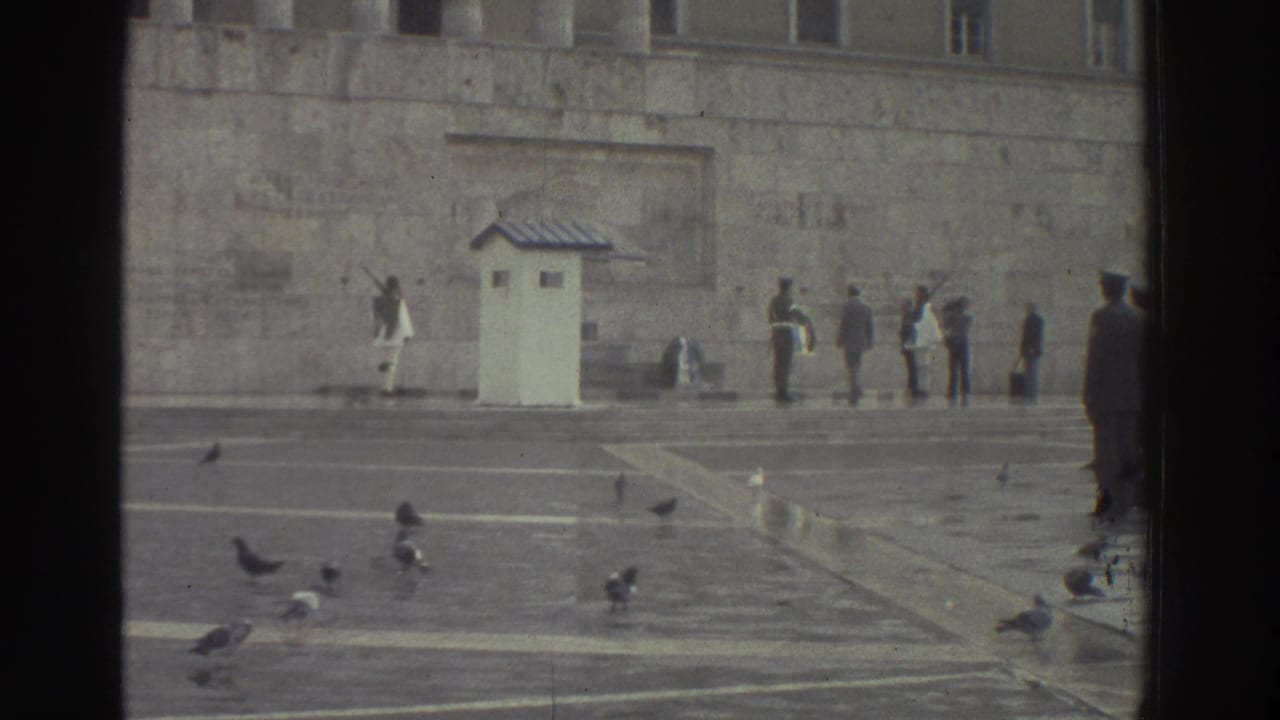 Wide shot of a stone plaza with a white guard booth, uniformed figures, civilian onlookers, and pigeons on wet paving in front of a large stone building.