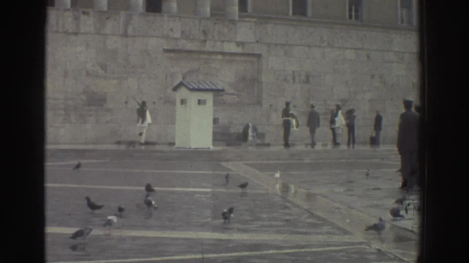 1976 Greece: stone public square with white sentry booth, white-uniformed guard, dark-uniformed personnel, civilian onlookers, pigeons on the pavement; large stone building behind.