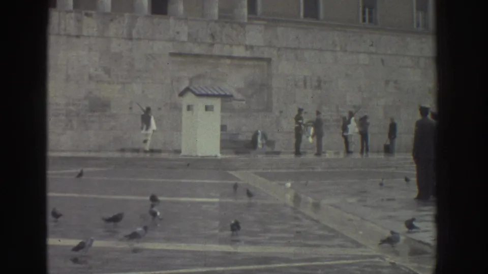 1976 Greece: wide shot of guard ceremony with sentry booth, white-clad guard, civilian group, and a silhouetted figure at right; pigeons scattered across the stone paving.