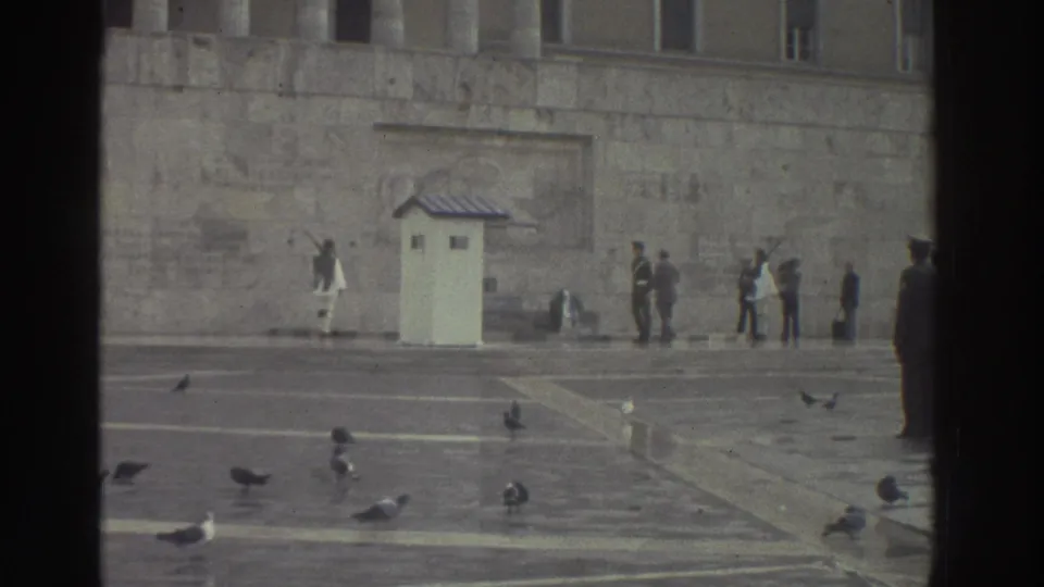 1976 Greece: stone ceremonial square with prominent foreground silhouette at right; white sentry booth and white-uniformed guard visible mid-frame; pigeons across the paving.