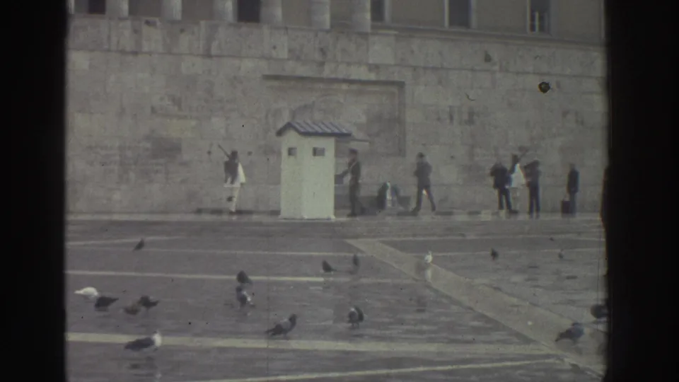 1976 Greece: closing frame of the ceremonial square with sentry booth, guards, civilian onlookers, and numerous pigeons; subdued overcast light across the stone surfaces.
