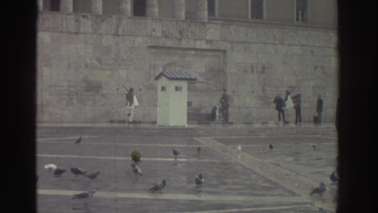 Wide shot of stone memorial monument with broad paved plaza; pigeons on steps; small guard booth at left; distant figures near building.