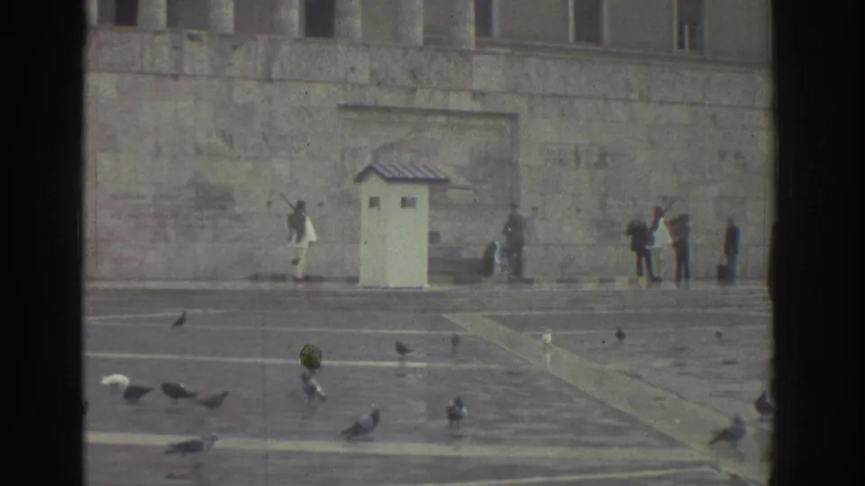 Wide establishing shot of stone memorial plaza in Greece, circa 1976; pigeons scattered on steps, guard booth visible at left, distant figures at monument.