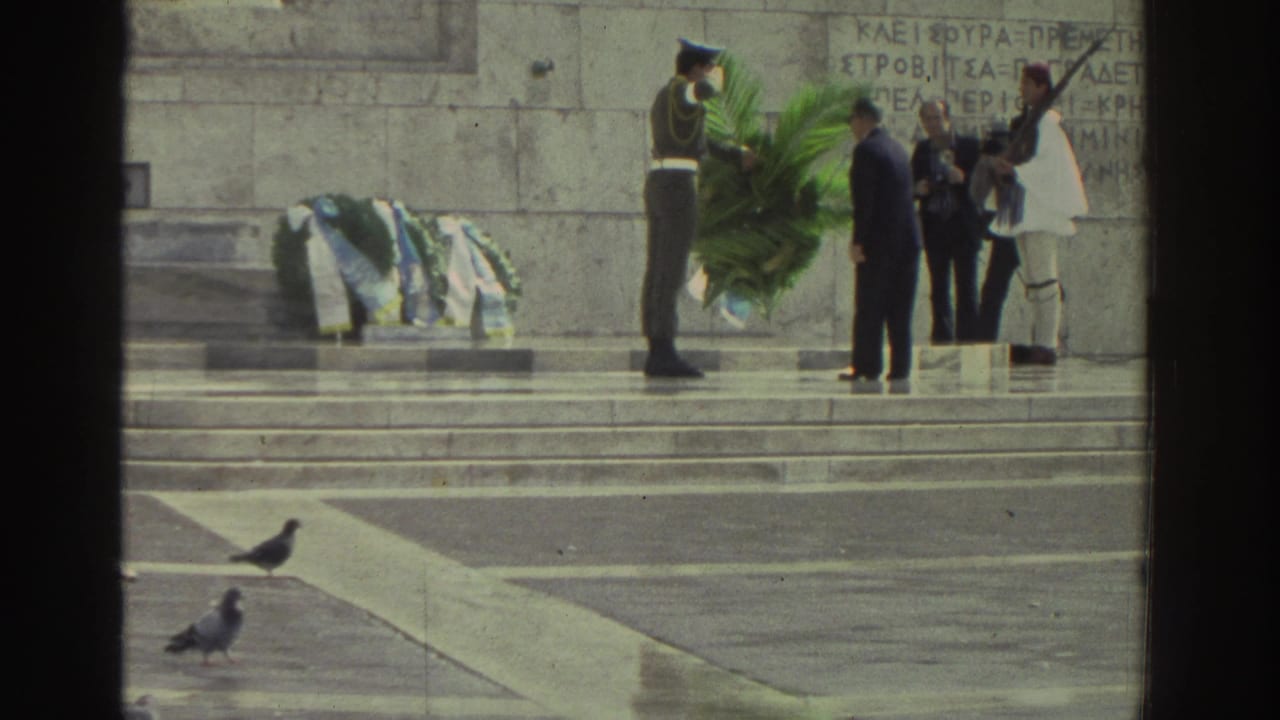 Closer view of Greek memorial steps; soldier and civilian figures with large green wreath; white-uniformed ceremonial guard visible; Greek text inscribed on stone wall.