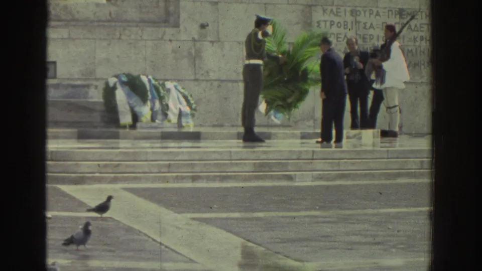 Wreath presentation at Greek memorial steps, circa 1976; uniformed soldier and civilian participants with green palm wreath; ceremonial guard in white visible; Greek inscriptions on stone.