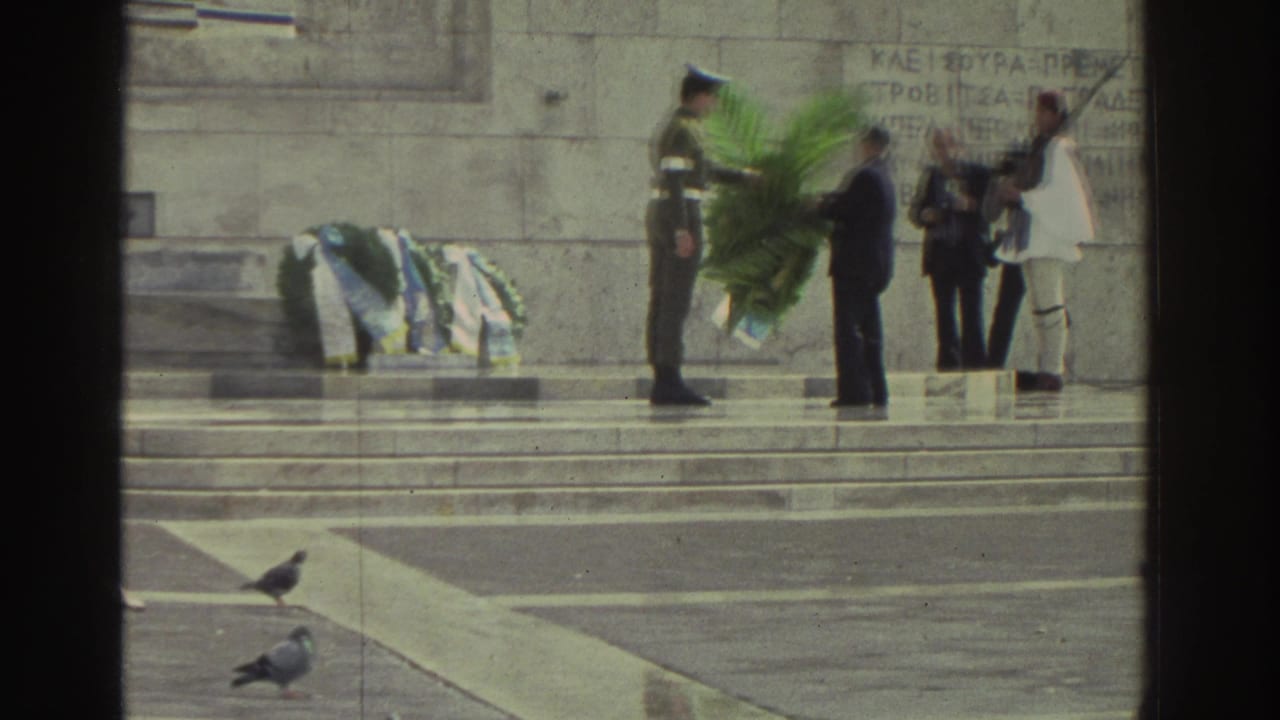 Wreath-laying ceremony at Greek memorial, circa 1976; soldier presenting large green palm-frond wreath to civilian; Evzone-style ceremonial guard in white at right; Greek text engraved on stone.
