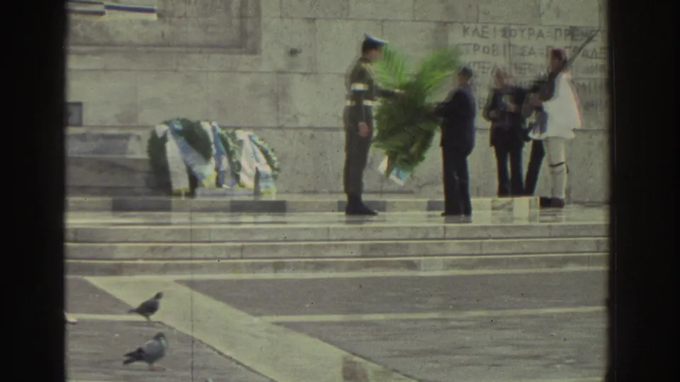 Wreath-laying ceremony at Greek memorial, circa 1976; soldier presenting large green palm-frond wreath to civilian; Evzone-style ceremonial guard in white at right; Greek text engraved on stone.