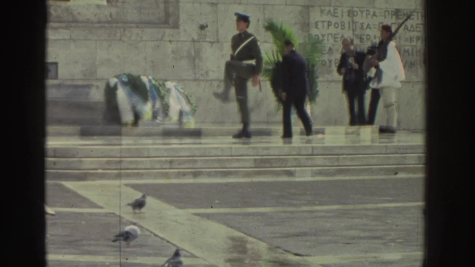 Greek memorial ceremony, circa 1976; ceremonial guard with rifle, civilian and uniformed participants, Evzone-style guards in white; blue-and-white wreaths laid on stone steps, pigeons below.