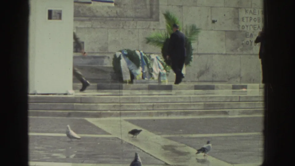Greece, 1976: figure in dark suit walks along stone monument steps; white guard booth and green wreaths visible below a wall bearing carved Greek text.