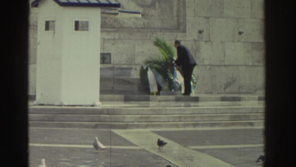 Greece, 1976: man in dark suit leans to place a large green wreath at the base of a stone national memorial; pigeons visible on the stone plaza in the foreground.