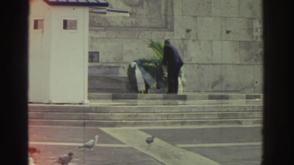 Greece, 1976: man in dark suit positions green wreath at the base of a stone national monument; pigeons on lower plaza steps; partial Greek text carved into stone wall visible upper right.