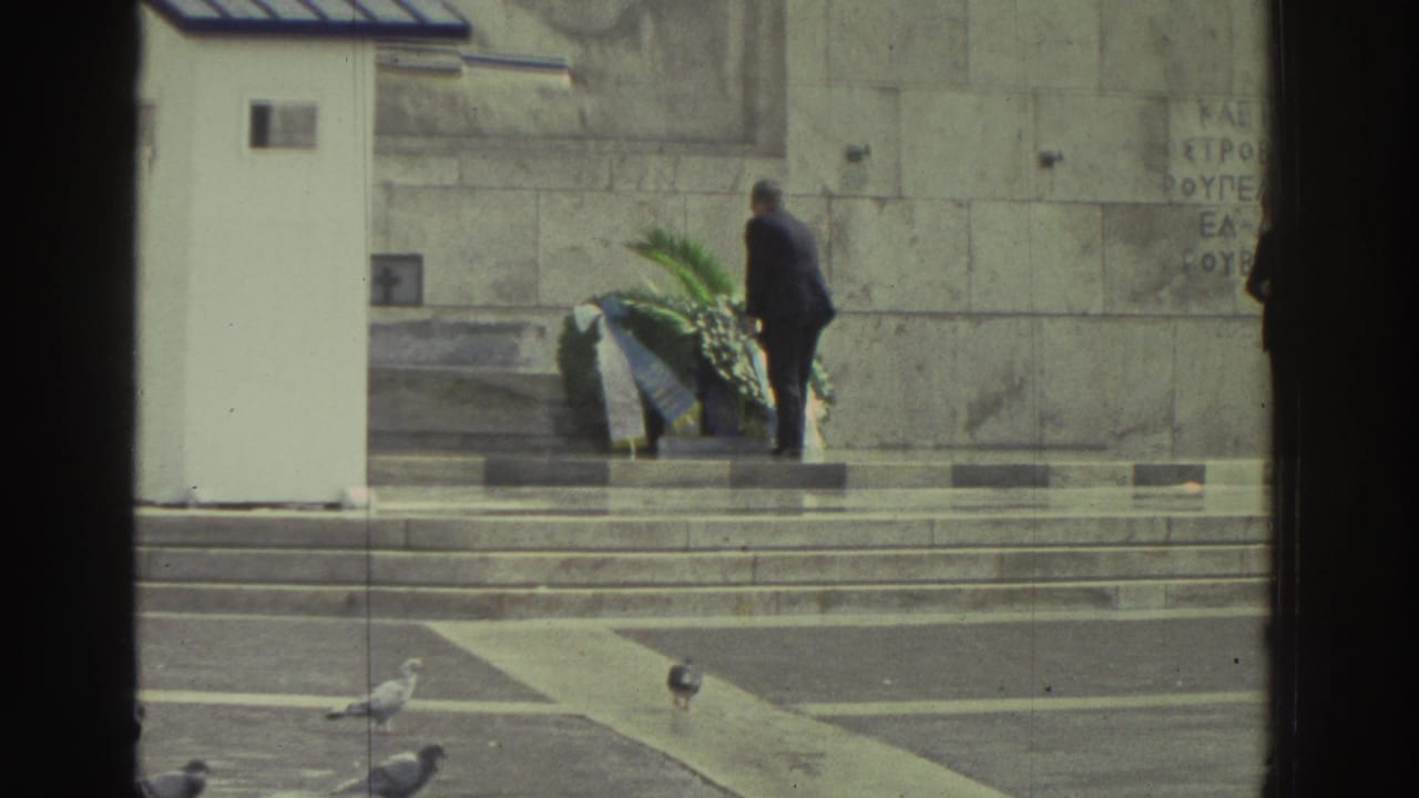 Man in dark coat stands beside a large floral wreath on steps of a stone memorial; pigeons on the paved forecourt below.