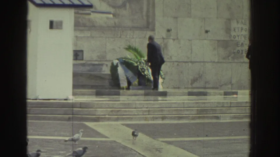 Lone figure placing or standing beside a large floral wreath at a stone memorial in Greece, circa 1976; pigeons on foreground pavement.