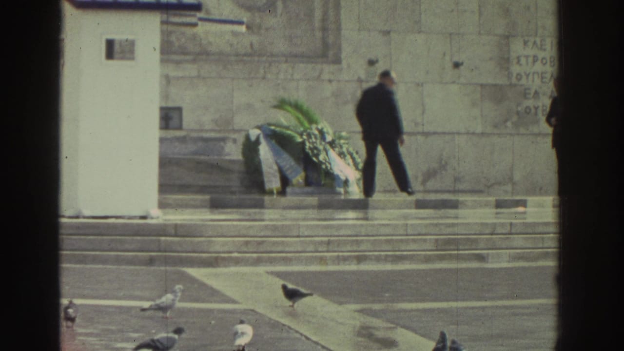 Stone memorial with several laid wreaths and departing figure; pigeons on forecourt; fragmentary Greek-script inscription visible on the facade; Greece, circa 1976.