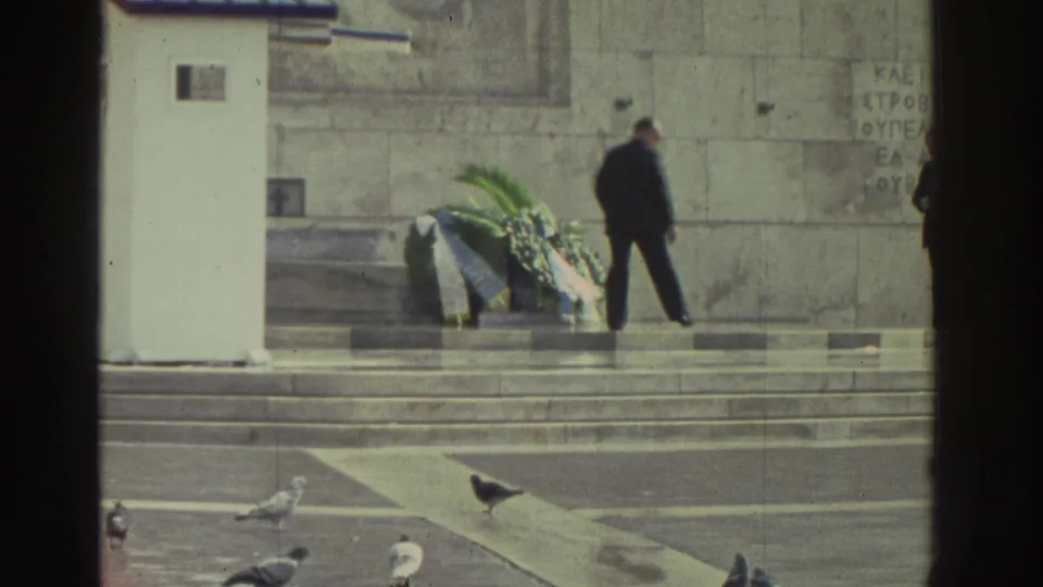 Stone memorial with several laid wreaths and departing figure; pigeons on forecourt; fragmentary Greek-script inscription visible on the facade; Greece, circa 1976.