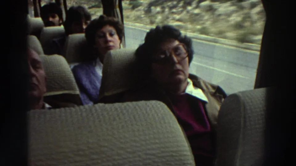 Coach interior in Greece circa 1976; woman in glasses and brown jacket seated in foreground, further passengers behind; rocky terrain through the window.