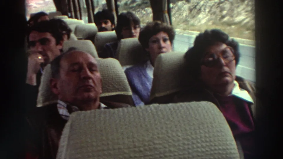 Candid coach interior, Greece circa 1976; older man dozing in foreground seat, several adult passengers visible mid-ground, rocky terrain through windows.