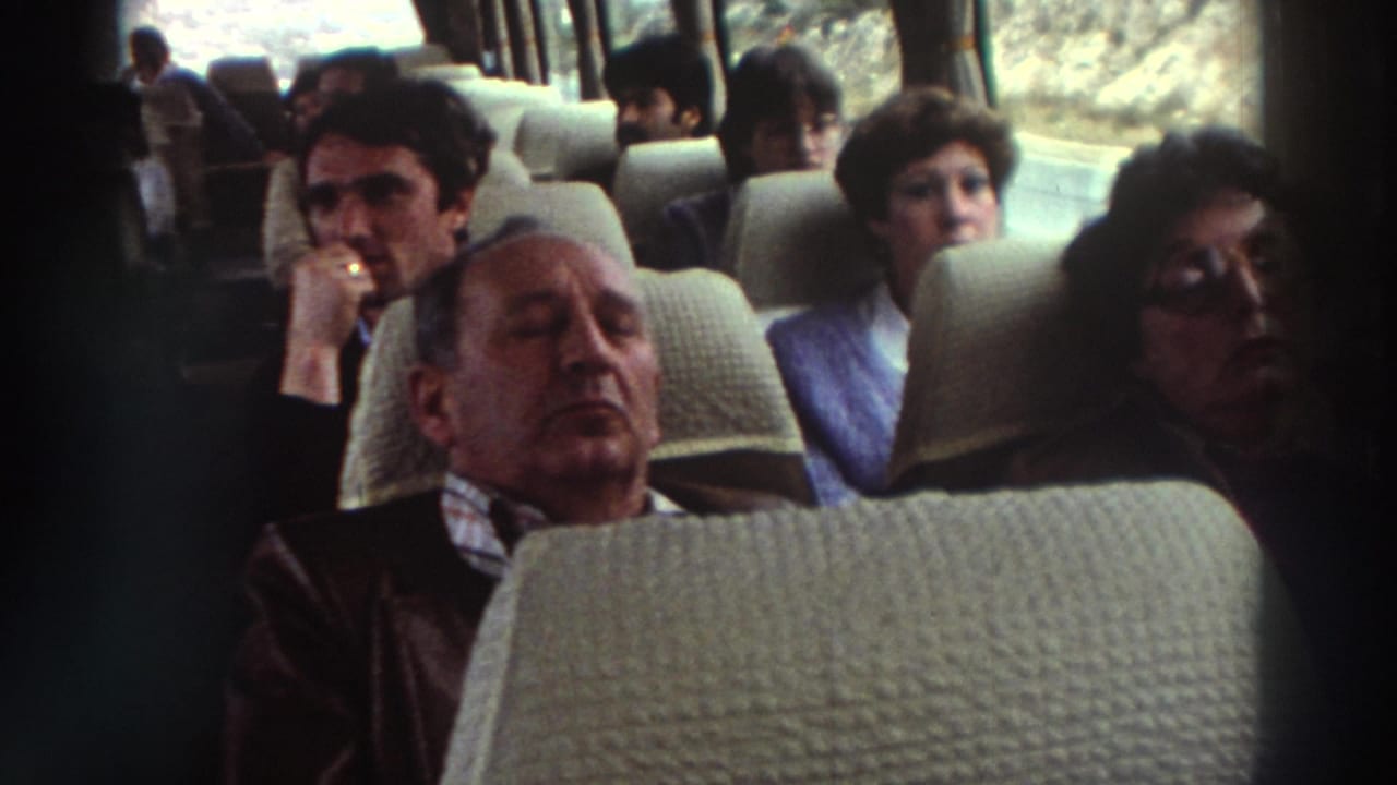 Older man asleep in a cream-upholstered bus seat during what appears to be a 1976 Greece coach tour, fellow passengers seated behind him.