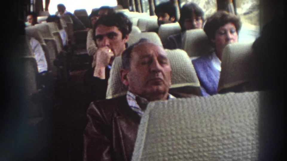 Continued view of a resting older man in a bus seat with fellow passengers visible behind him, consistent with a 1970s Greece group tour.