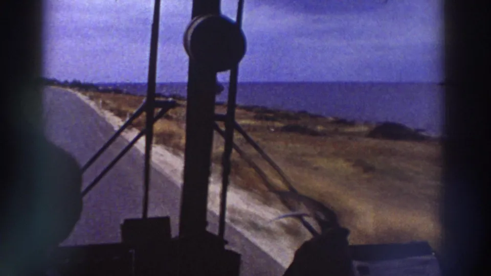 View from inside a bus showing a winding coastal road with arid terrain and a blue sea horizon, consistent with a 1970s Mediterranean road trip.