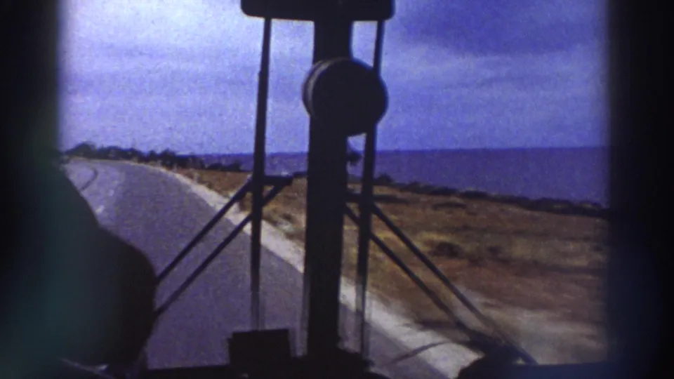 Continuing bus windshield view of a coastal road with blue sea and overcast sky, consistent with mid-1970s Mediterranean travel footage.