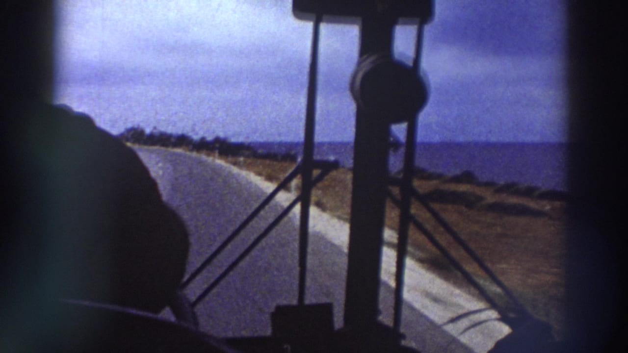 View through a bus window and metal seat rail toward a winding coastal road, sea visible on the horizon under a cloudy blue sky.