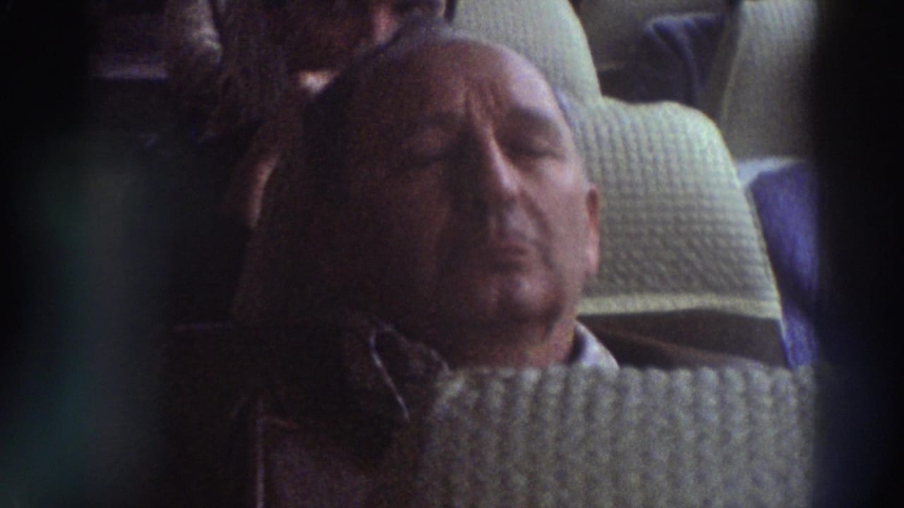Older balding man with eyes closed dozing in a quilted bus seat, wearing a light cable-knit sweater; second passenger partially visible behind.