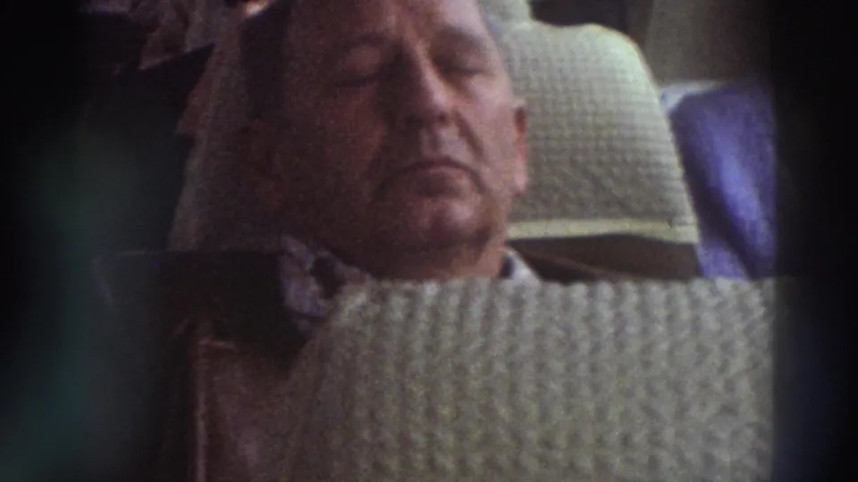 8mm close-up of an older male tourist dozing on a Greek coach, circa 1976; quilted seat fabric and cable-knit sweater prominent.