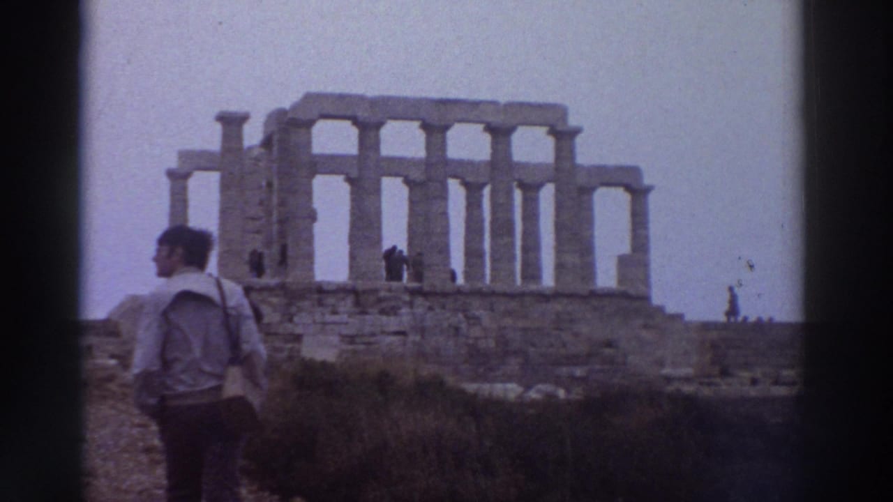 Man in light jacket standing with back to camera before ancient stone temple ruins with tall standing columns.