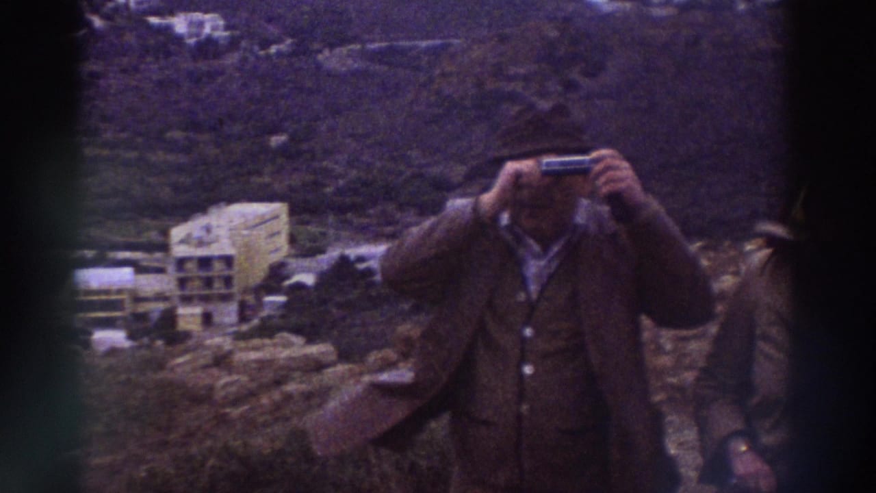 Man in dark jacket raising binoculars to his face, with a coastal hillside and large concrete building visible behind him.