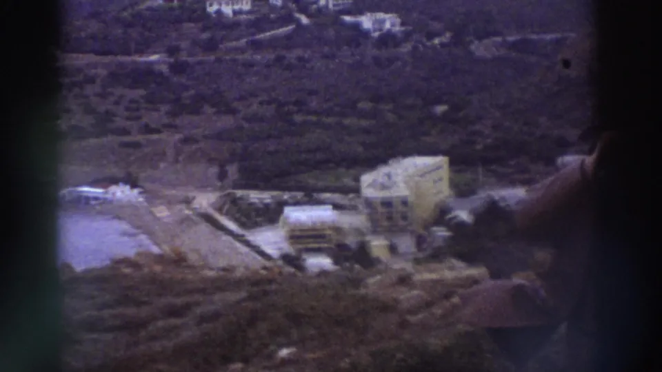 Elevated view of a Greek coastal area circa 1976 with a large modern building, shoreline, scrub hillside, and a hand pointing into frame, 8mm home movie.