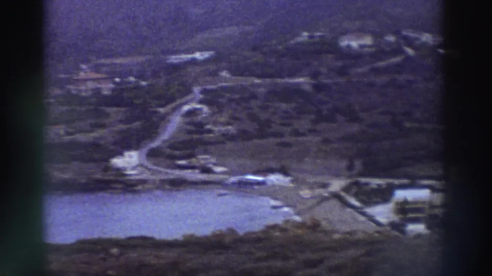 Panoramic elevated shot of a Greek coastal area circa 1976 showing blue sea, a winding road, and sparse white buildings on scrubby hillside, 8mm home movie.