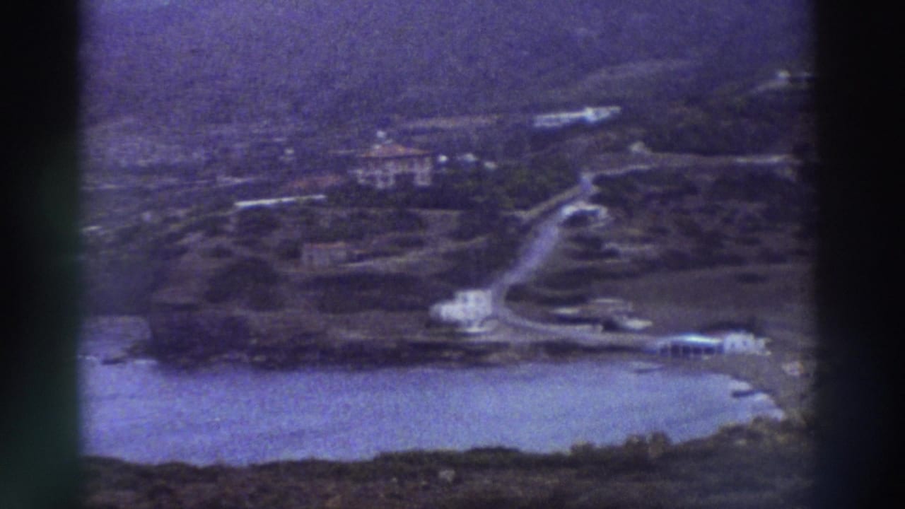 1976 8mm home-movie frame: elevated view of a small Greek coastal settlement with whitewashed buildings, a sheltered bay, and a winding hillside road.