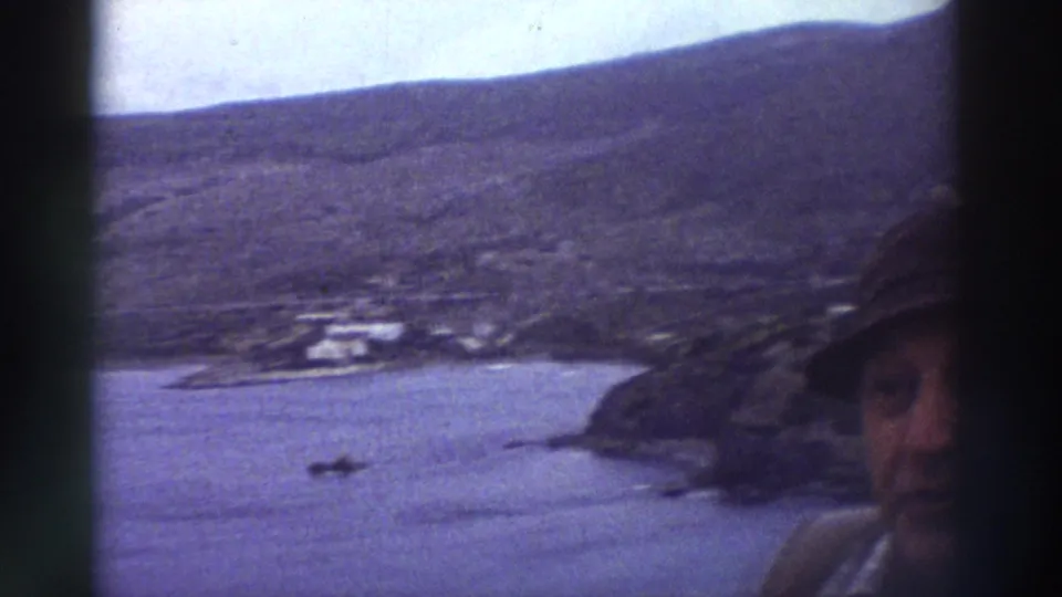 1976 8mm home-movie frame: rocky Greek headland and small harbour from an elevated angle; a person's face is partially visible at the right frame edge.