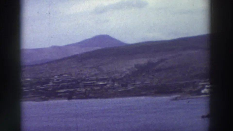 1976 8mm home-movie frame: wide panoramic view of a hazy Greek mountain range above a coastal settlement and calm sea.