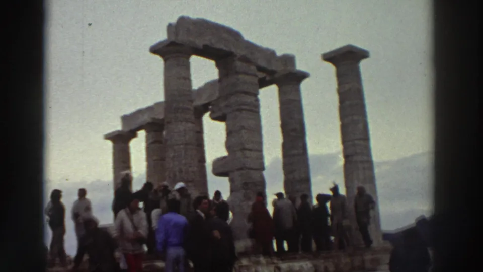 1976 8mm home-movie frame: crowd of tourists at the base of standing Doric columns at a Greek ancient ruin, appearing to overlook the sea.