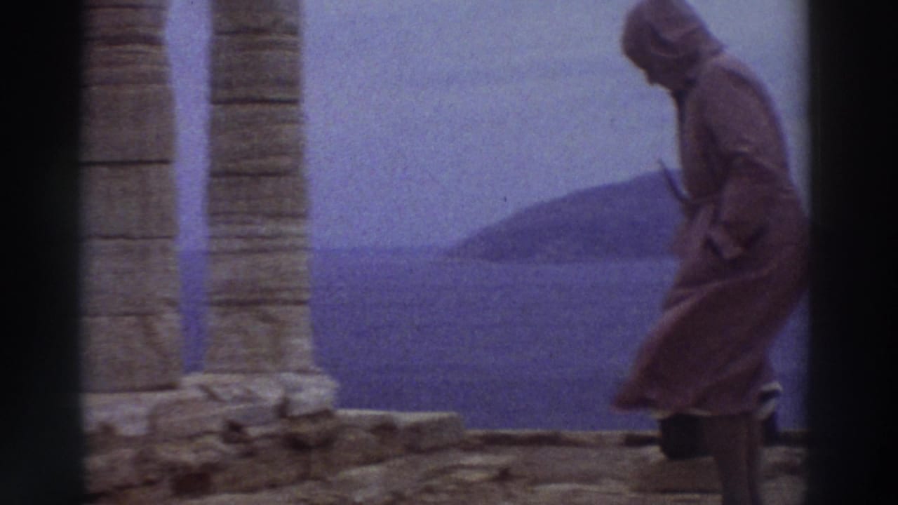 A hooded visitor walks among tall fluted stone columns at an ancient Greek site in 1976, with a coastal headland visible across the sea in the background.