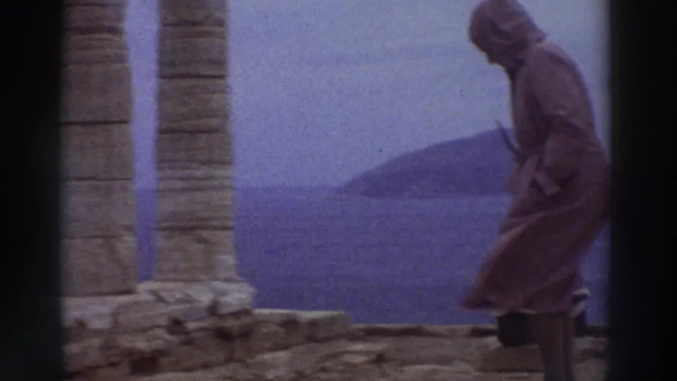 A hooded visitor walks among tall fluted stone columns at an ancient Greek site in 1976, with a coastal headland visible across the sea in the background.