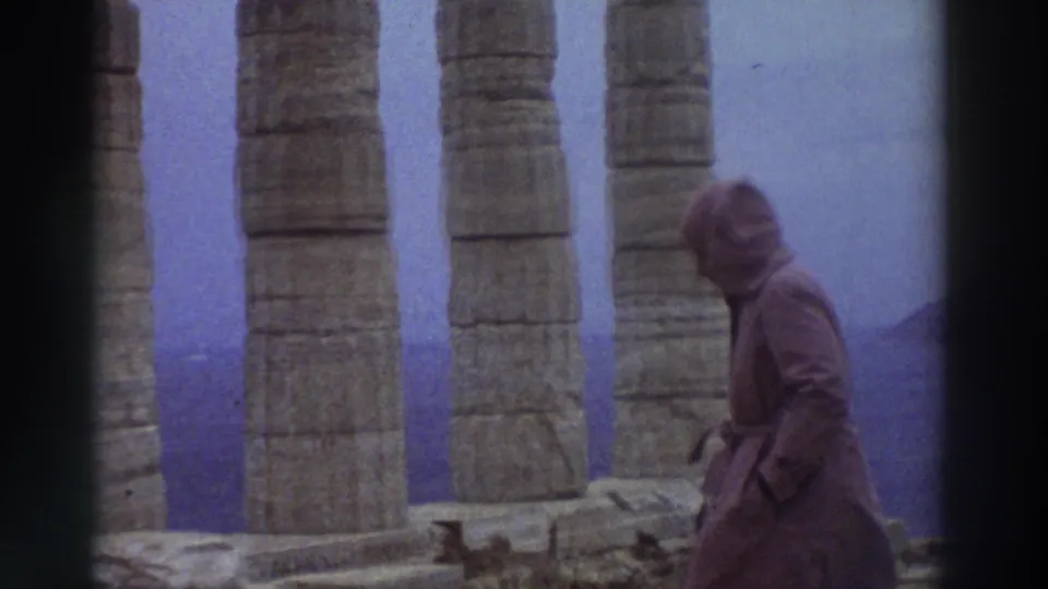 A hooded visitor begins to turn among ancient Greek stone columns in 1976, with a distant dark coastal landmass visible through gaps in the colonnade.