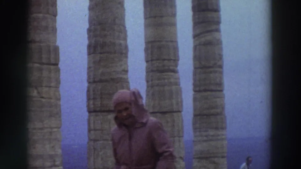 A hooded visitor faces the camera at an ancient Greek columned site in 1976, partial face visible under the hood, flanked by tall stone columns.