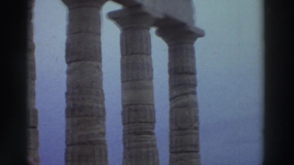 Ancient stone columns at a Greek archaeological site in 1976 fill the frame against a pale sky, with the hooded figure partially visible at the lower right.