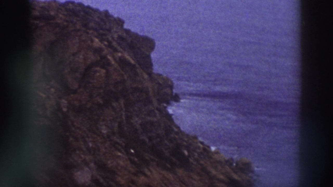 Steep reddish-brown rocky cliff descending to purple-blue sea, 8mm with visible color drift.