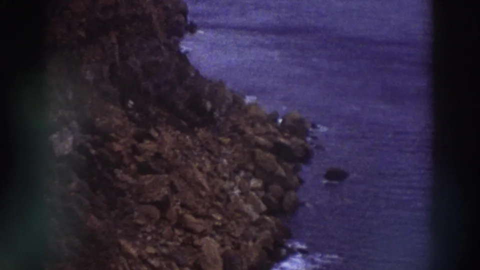 Rocky Greek coastline with scattered boulders and calm sea filmed on 8mm circa 1976, showing purple color cast from aged film stock.