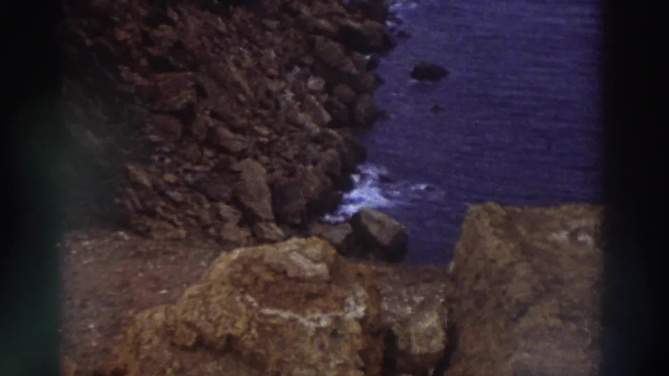 Textured rocky foreground with sea and white surf behind on Greek coastline circa 1976, filmed on 8mm with warm ochre tones in foreground rocks.