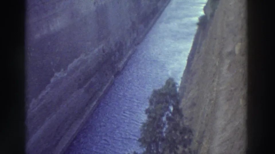 8mm home movie from Greece, c.1976: continuation of narrow canal shot, steep stone-cut walls, blue-purple water, tree on embankment, lavender color cast.