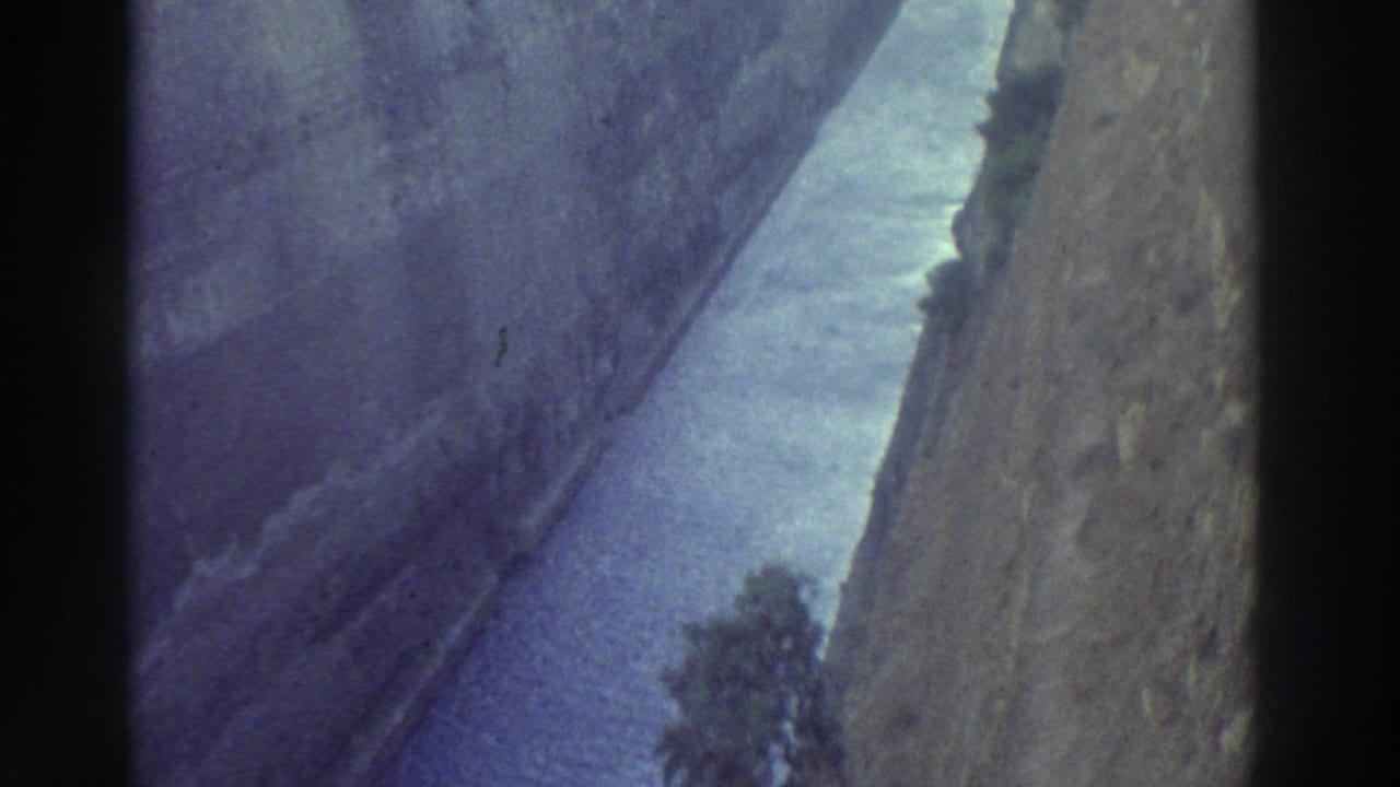 1970s 8mm shot from the rim of a narrow canal cut through steep rock walls, blue-purple water below with small trees at the base, Greece 1976.