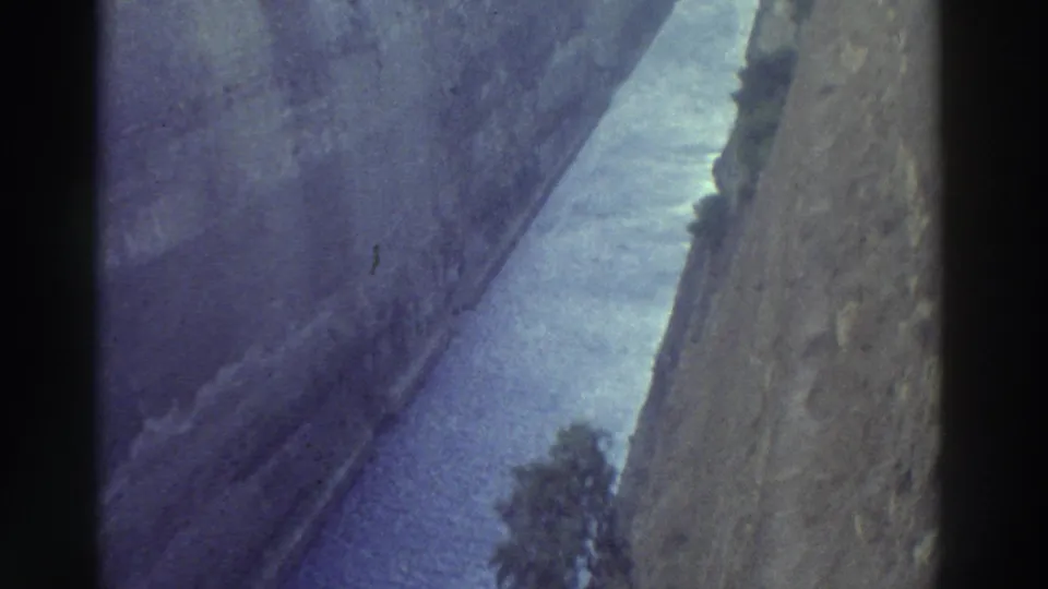 1970s 8mm shot from the rim of a narrow canal cut through steep rock walls, blue-purple water below with small trees at the base, Greece 1976.
